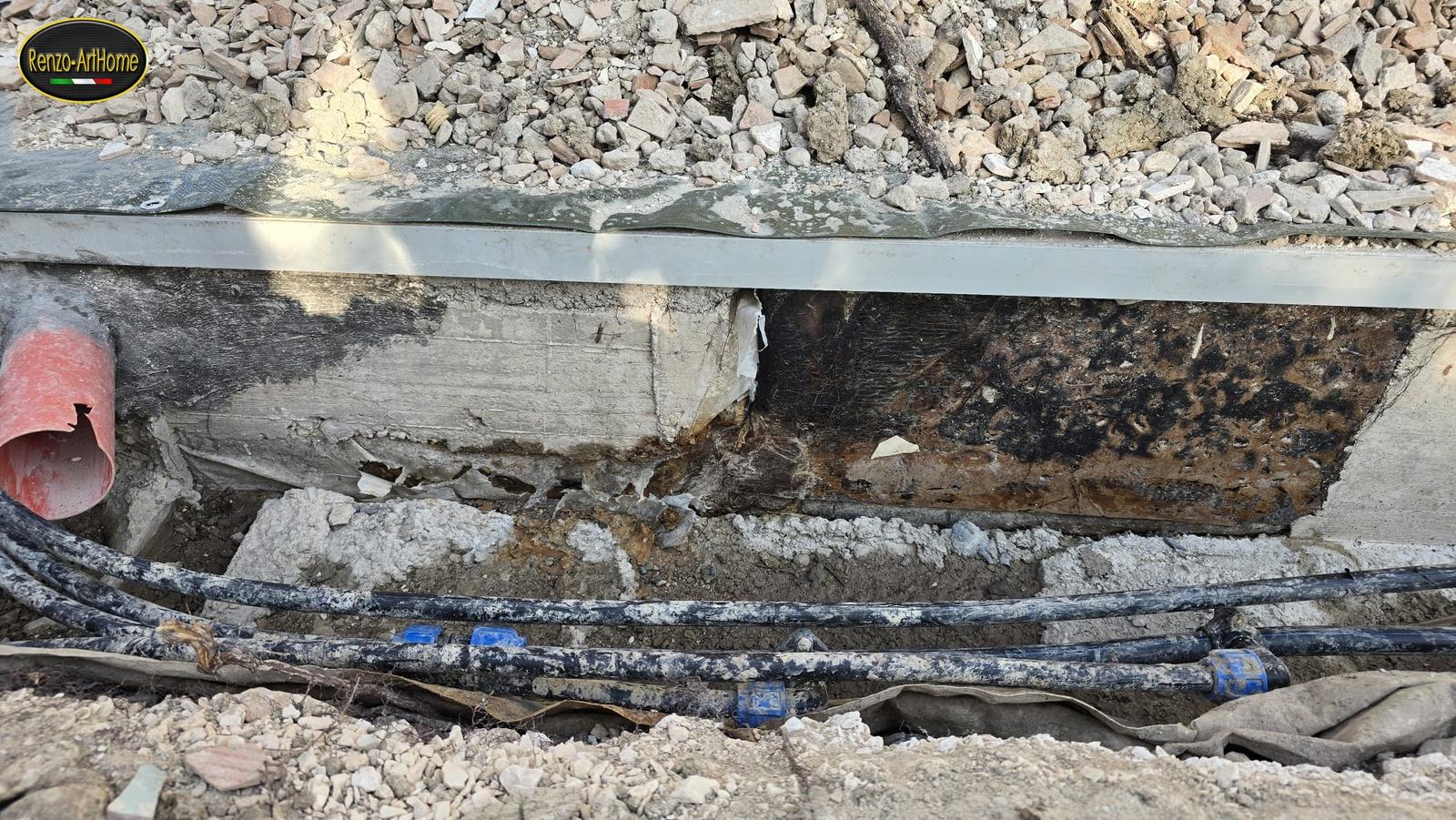Base di un muro messa a nudo dallo scavo, con la vecchia spalmatura di bitume ridotta a croste scure e staccate, e i cavi degli impianti che corrono lungo lo scavo