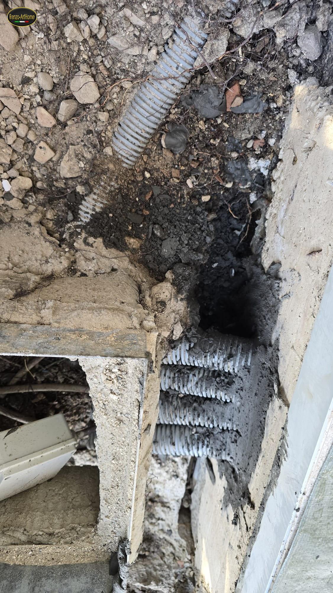 Muro interrato con i tubi corrugati degli impianti che lo attraversano, il vuoto scuro intorno ai tubi e la muratura tagliata per farli passare