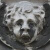 Testa d'angelo in pietra con le ali, su una tomba del cimitero del Père-Lachaise a Parigi: nere le parti riparate sotto gli occhi, intorno al naso e nell'incavo delle ali, chiare le parti in rilievo lavate dalla pioggia