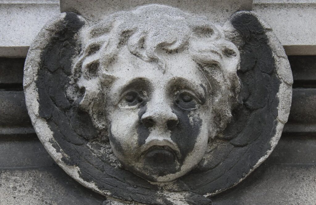 Testa d'angelo in pietra con le ali, su una tomba del cimitero del Père-Lachaise a Parigi: nere le parti riparate sotto gli occhi, intorno al naso e nell'incavo delle ali, chiare le parti in rilievo lavate dalla pioggia