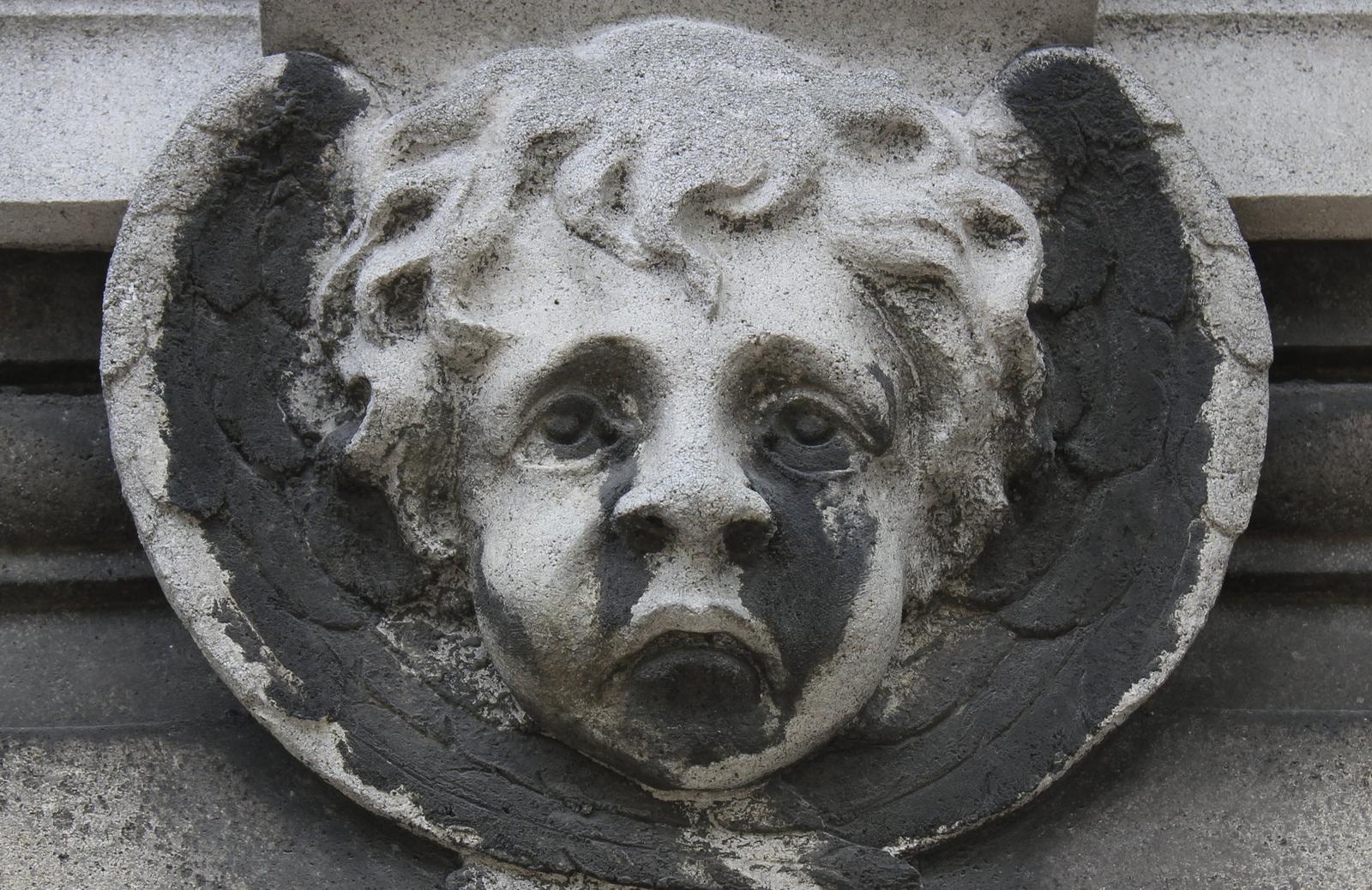 Testa d'angelo in pietra con le ali, su una tomba del cimitero del Père-Lachaise a Parigi: nere le parti riparate sotto gli occhi, intorno al naso e nell'incavo delle ali, chiare le parti in rilievo lavate dalla pioggia