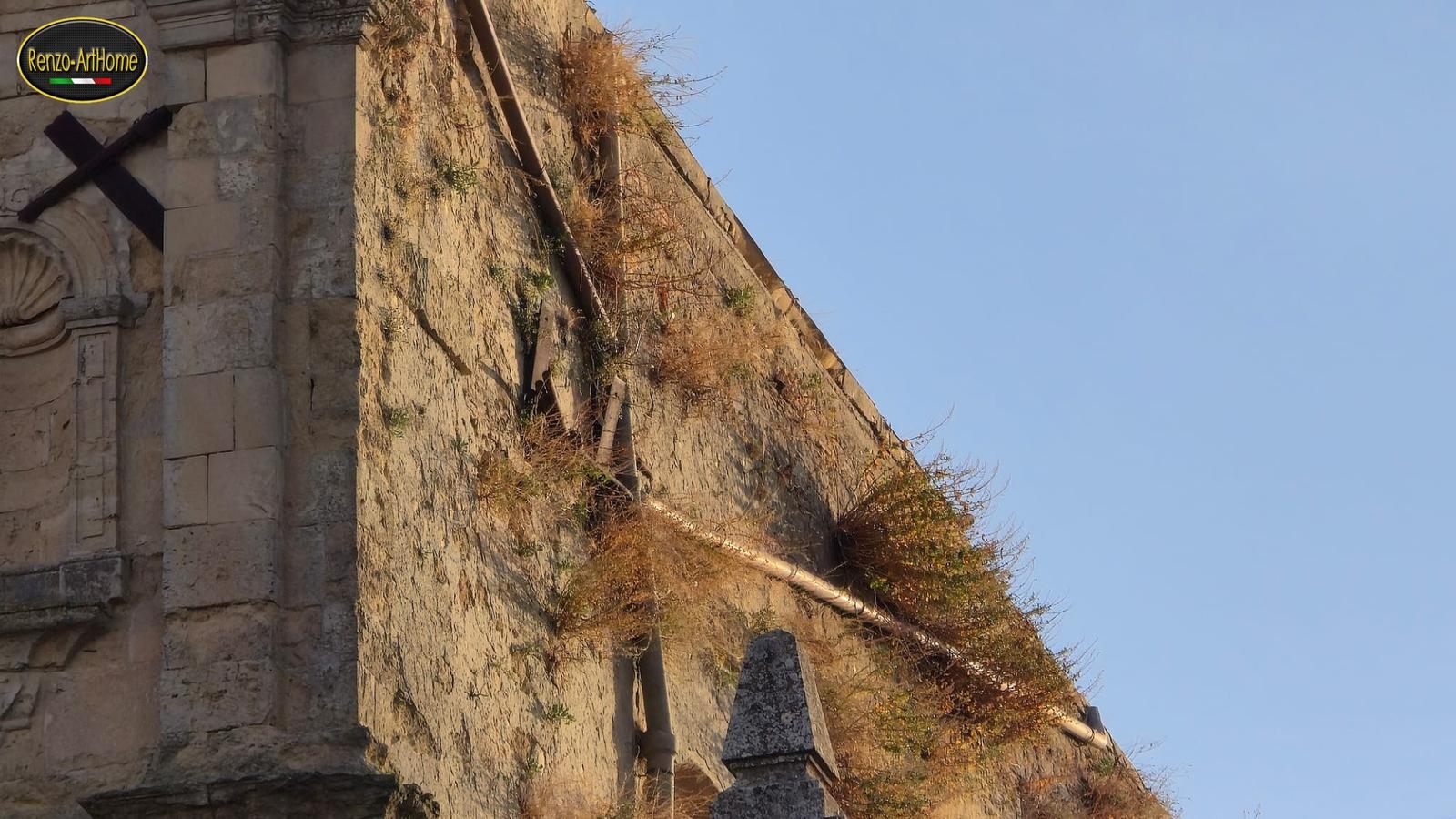 Il fianco in pietra di una chiesa visto dall'angolo della facciata, con i cespugli secchi e verdi che crescono lungo il bordo alto del muro e lungo il tubo di scarico, rotto a metà con gli spezzoni staccati dal muro; a sinistra i conci squadrati della facciata, con una nicchia a conchiglia e una croce