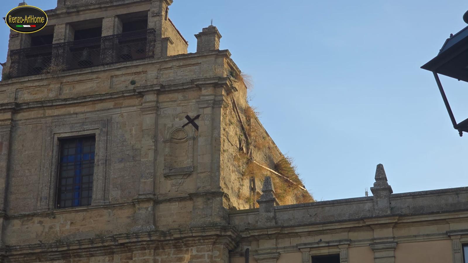 La chiesa della Fotografia 1 ripresa da più lontano: in alto la cella campanaria con le campane dietro la ringhiera, sotto la facciata in conci squadrati con le lesene, la finestra, la nicchia a conchiglia e la croce, quasi senza verde; a destra il fianco, preso dal sole, con i cespugli lungo il bordo alto del muro e lungo il tubo rotto; in basso a destra il parapetto con i pinnacoli di un edificio più basso